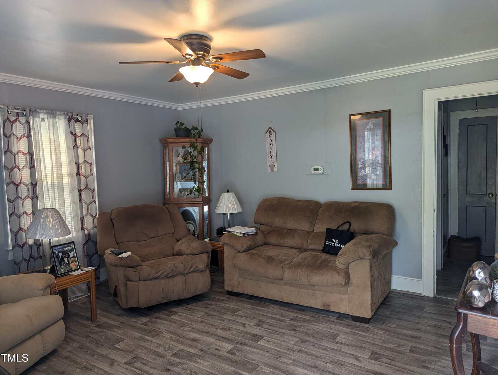 A cozy living room with gray walls and wood plank flooring. It features a brown suede sofa, matching love seat, and recliner. There's a wooden side table with a lamp, a ceiling fan with light, and large windows with patterned curtains. Decor includes framed pictures.