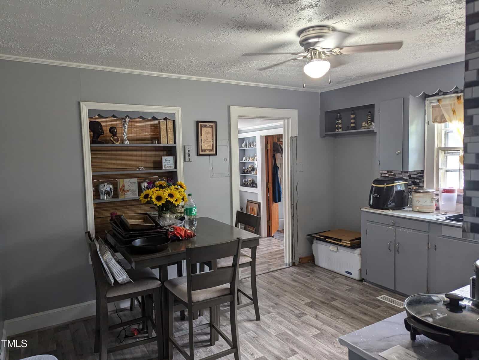 A kitchen with gray cabinets and walls, featuring a ceiling fan with a light. A dining table with four chairs has a laptop and sunflowers on it. A window with a curtain allows natural light, and various kitchen appliances are on the countertops. Floor is wood-look vinyl.