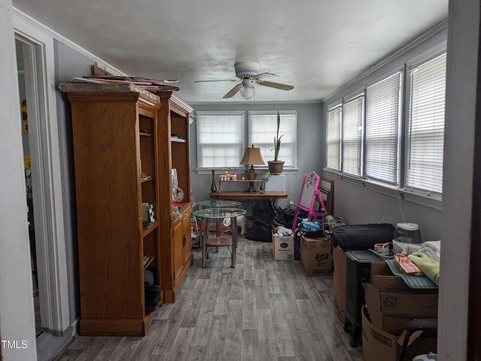 A small room cluttered with various items including boxes, bags, and a pink children's chair. The room has large windows with blinds, a ceiling fan, wooden shelves on the left, and a glass-top table in the center. The floor is covered with gray wood-like tiles.