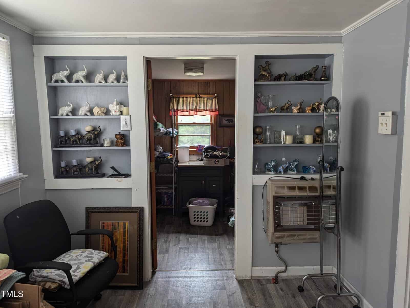 A small room with light blue walls, two built-in shelves filled with figurines on either side of an opening to another room with wood-paneled walls. The second room has a hamper and window. An office chair and some framed pictures are on the left, and an electric heater is on the right.