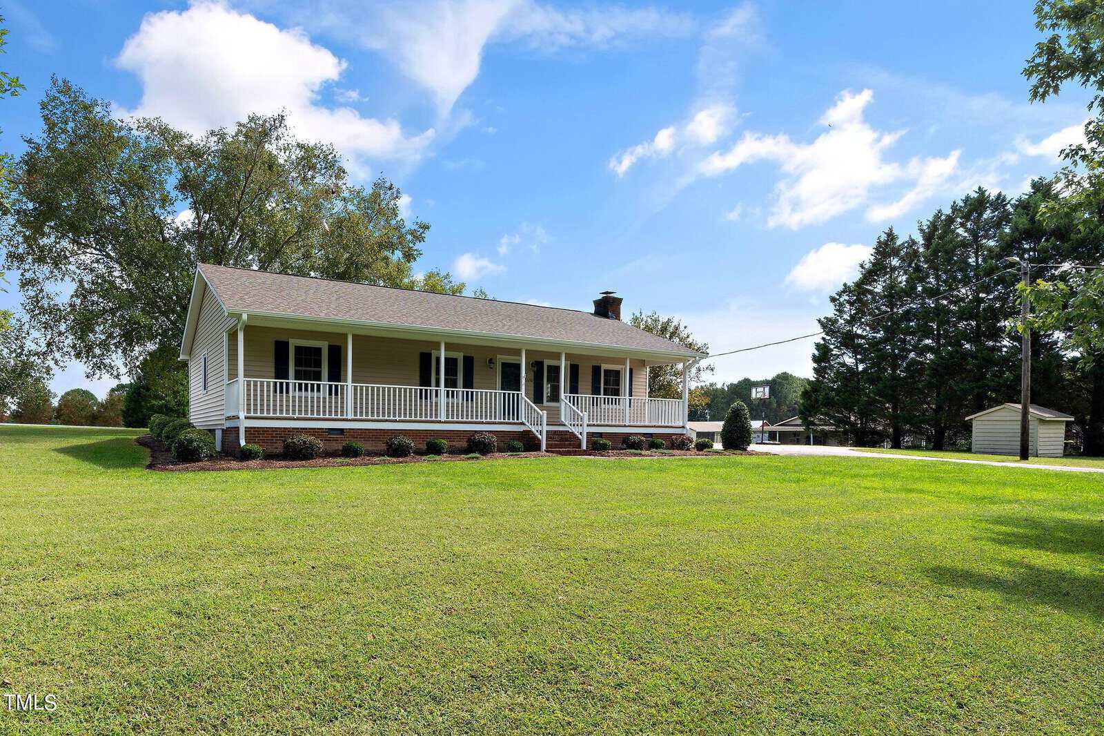 A charming single-story house at 1678 Hayes Road in Creedmoor, featuring a spacious front porch and surrounded by a well-maintained lawn. The sky is clear with a few clouds, and trees encircle the property. A long driveway leads to a small shed in the background.
