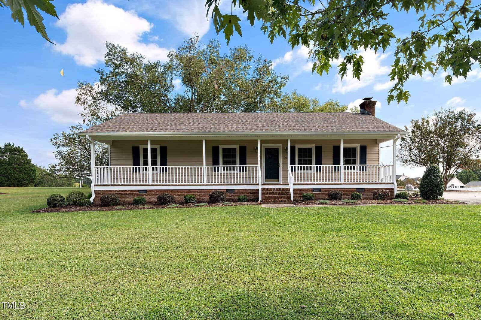 A beige, single-story house with a large front porch, white railings, and a chimney, surrounded by a well-maintained lawn and trees under a blue sky with some clouds.