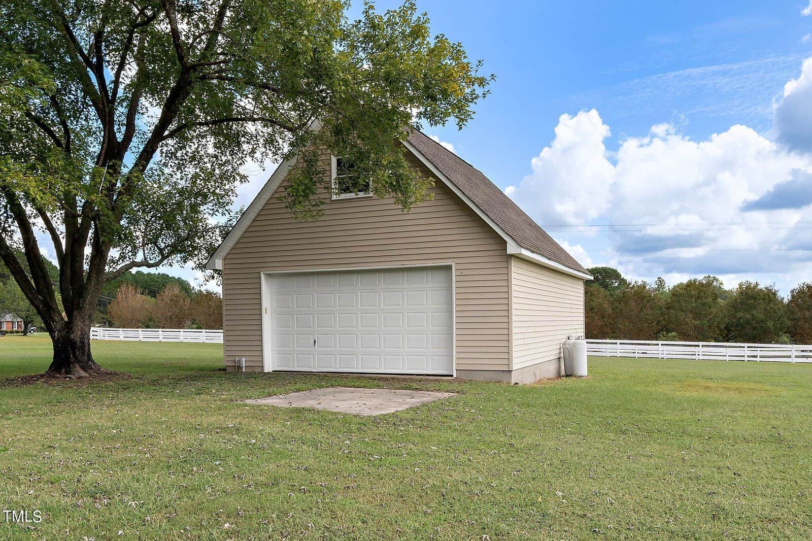 A small beige garage with a white door, surrounded by a grassy lawn and trees. The sky is partially cloudy, and there's a white fence in the background.