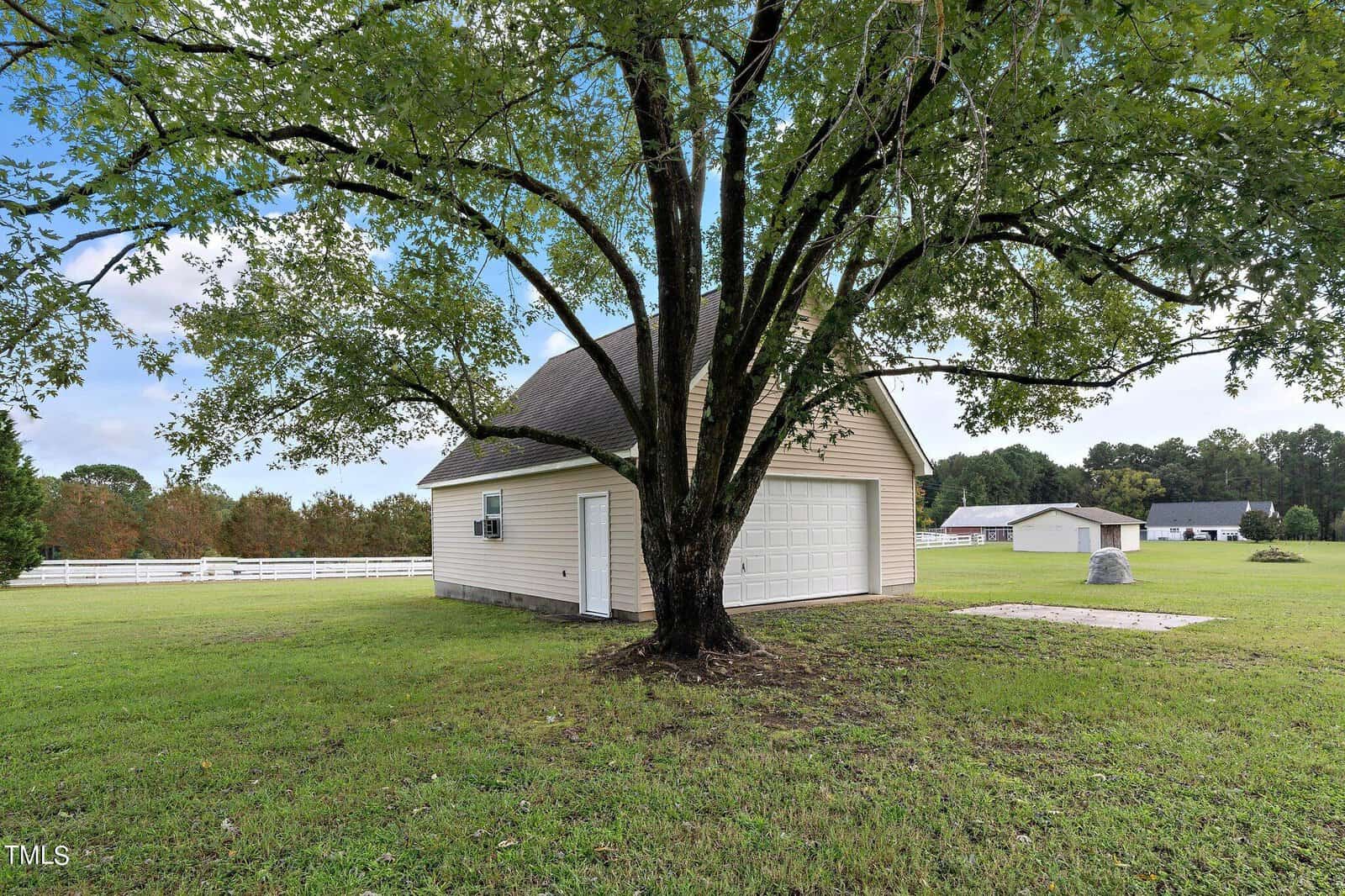 A white garage with a sloped roof stands under a large leafy tree in a grassy area. In the background, there are fenced fields, a distant small white building, and a house, all under a partly cloudy sky.