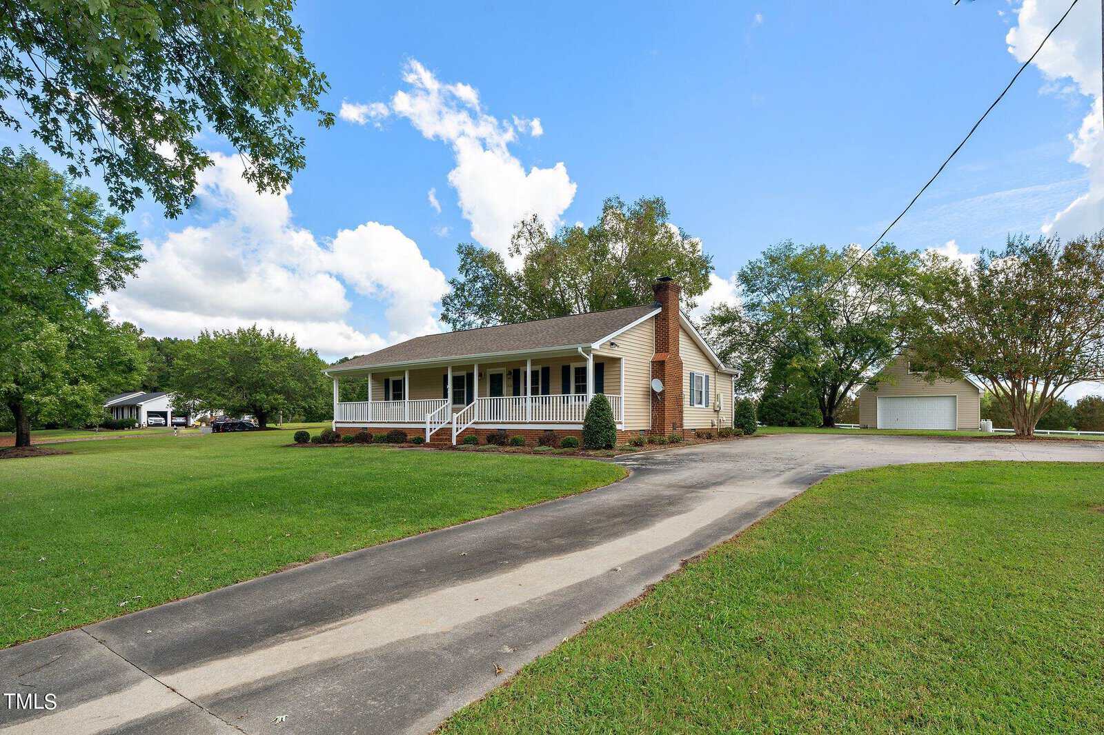 A single-story brick house with a front porch sits surrounded by a large green lawn. A winding paved driveway leads to a detached garage. The sky is blue with fluffy clouds, and tall trees frame the scene.