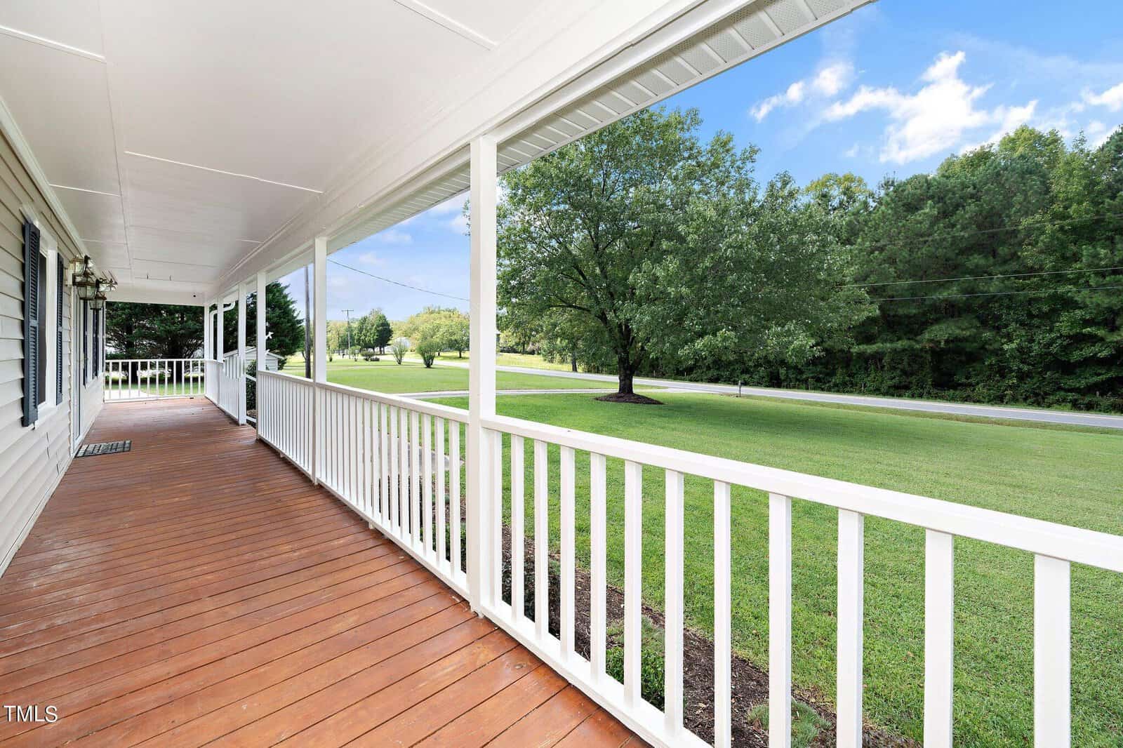 A spacious wooden porch with white railings overlooks a lush green yard and a tree-lined road under a blue sky with scattered clouds.