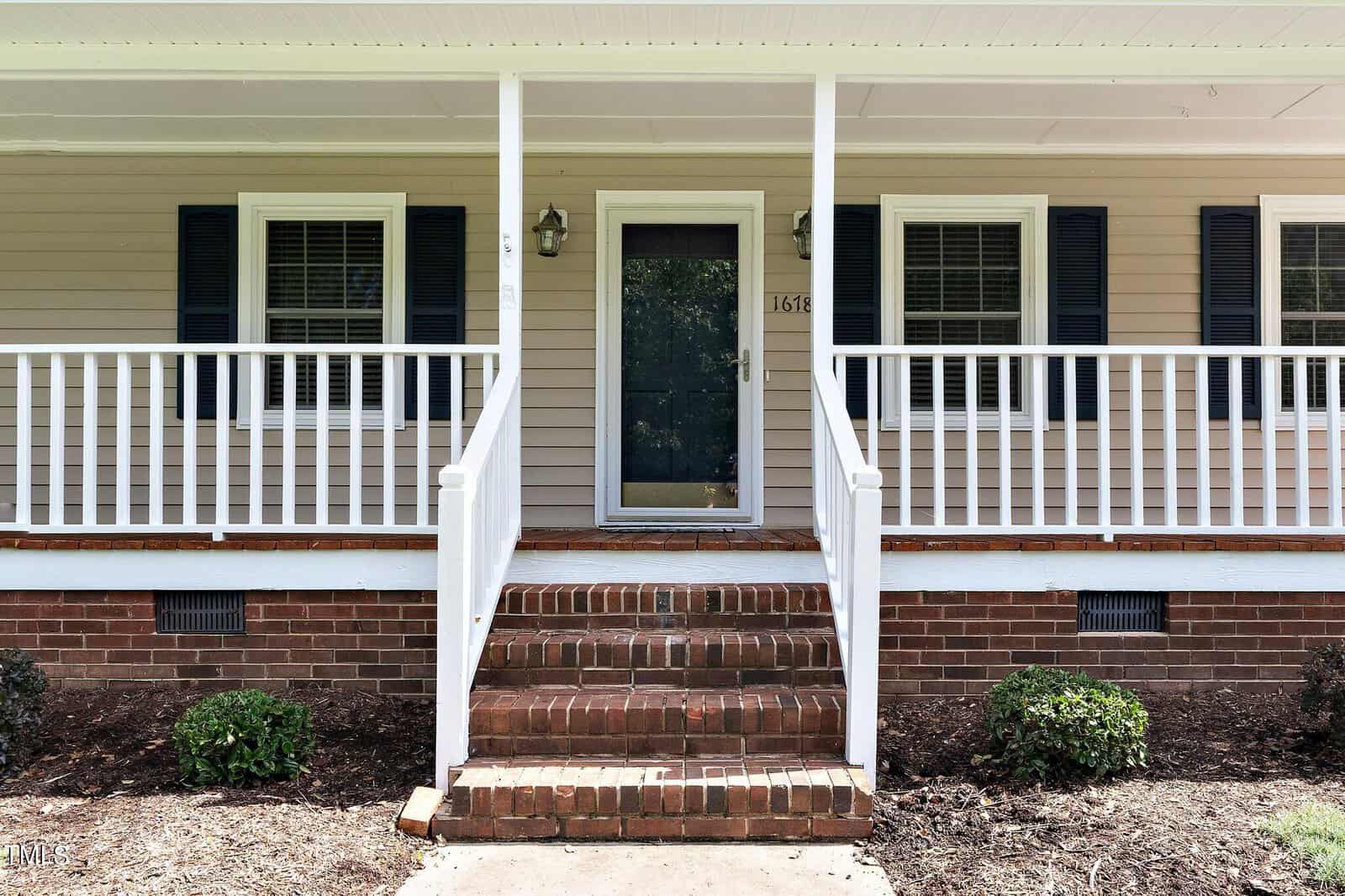 Front view of a house featuring a small porch with white railings and steps leading up to a central door. The exterior is light-colored siding with two windows on either side of the door, each with dark shutters. Two small shrubs are in front.