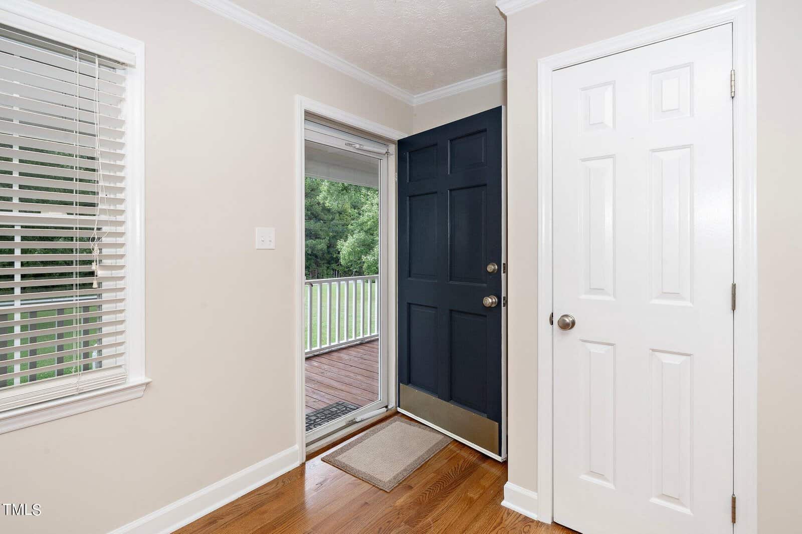A bright entryway with a dark blue front door open to a wooden deck outside. The room has cream-colored walls, white trim, a white interior door, and a window with white blinds to the left. Natural light illuminates the hardwood floor.