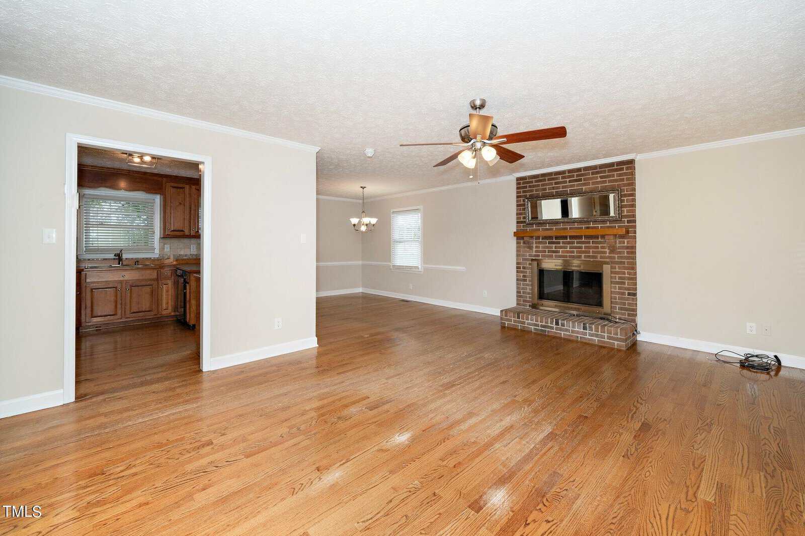 Spacious living room with polished wood flooring, a ceiling fan, and a brick fireplace with a mirror above it. The room opens to a kitchen with wooden cabinets. Neutral-colored walls and large windows provide ample natural light.