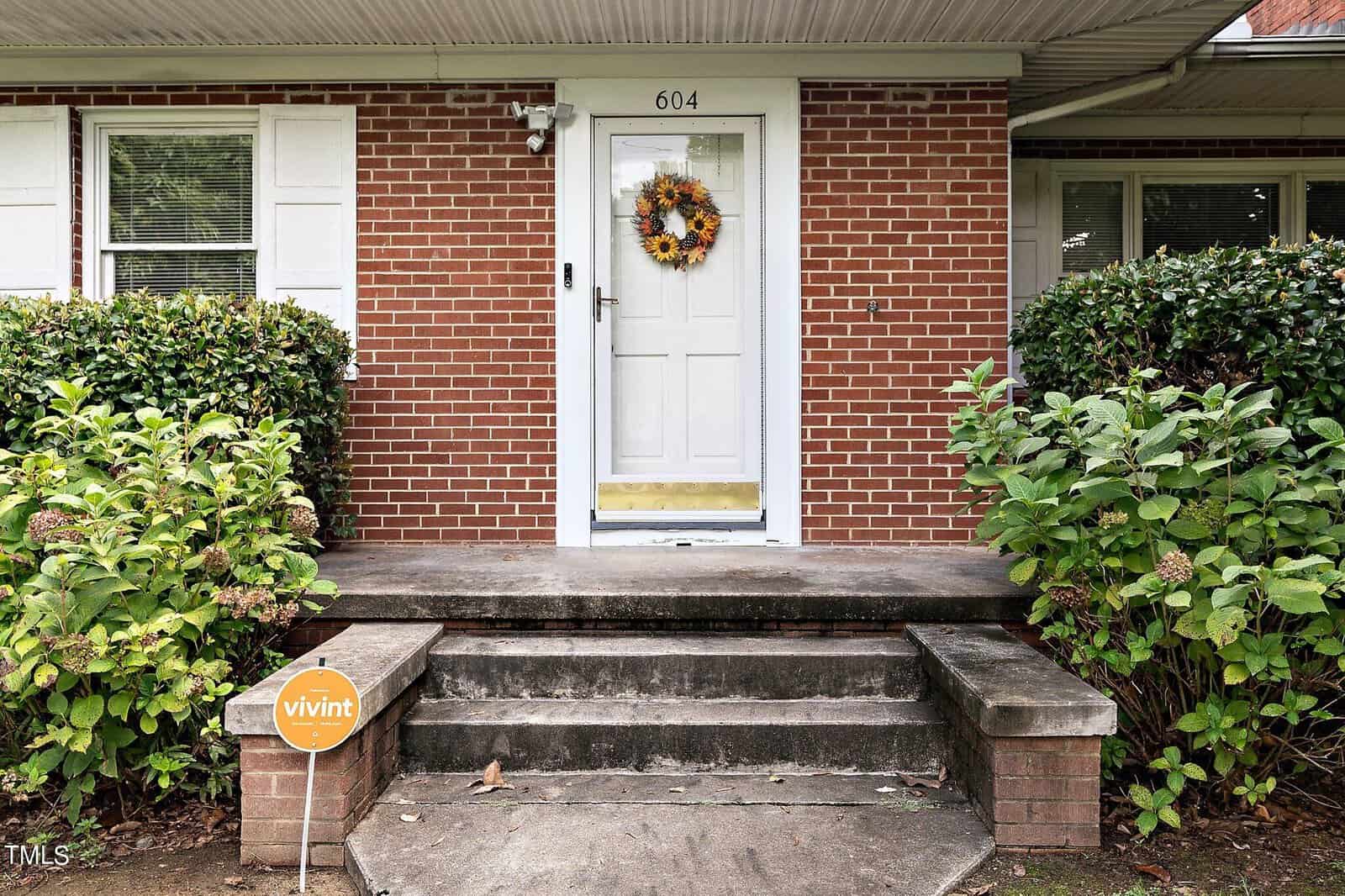 A white front door with a sunflower wreath is centered on a brick facade, flanked by green shrubs and white shutters. Three concrete steps lead up to the porch. A small Vivint security sign is staked in the grass to the left.
