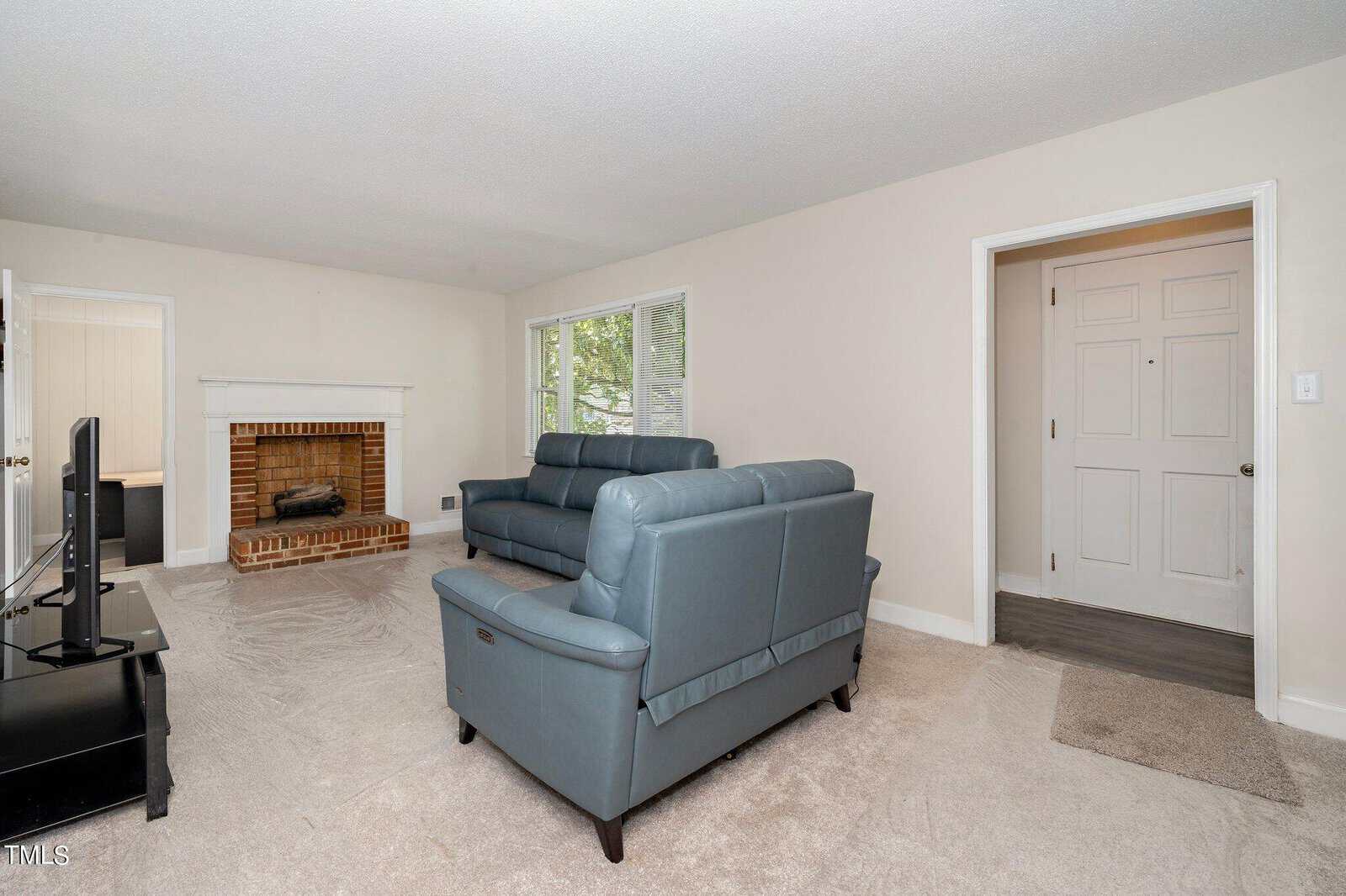 A living room with white walls and carpeted floor features a brick fireplace, two blue sofas, a black coffee table, and a television. A large window lets in natural light. The room leads to an entryway with a white door.