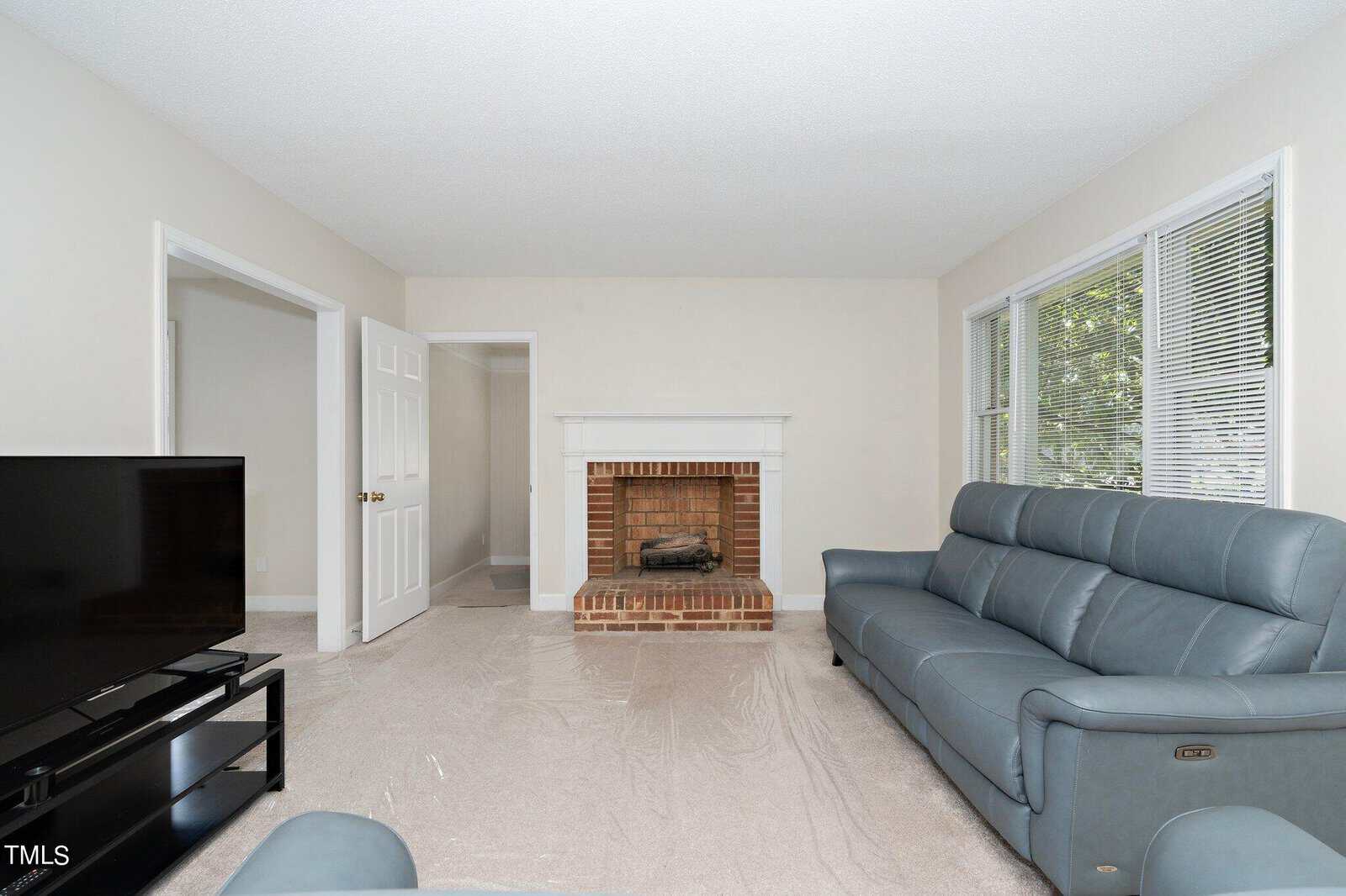 A living room with light gray walls featuring a brick fireplace and a white mantle. There are two blue leather sofas facing a large flat-screen TV on a black stand. The floor is covered with a transparent protective sheet. A window on the right lets in natural light.
