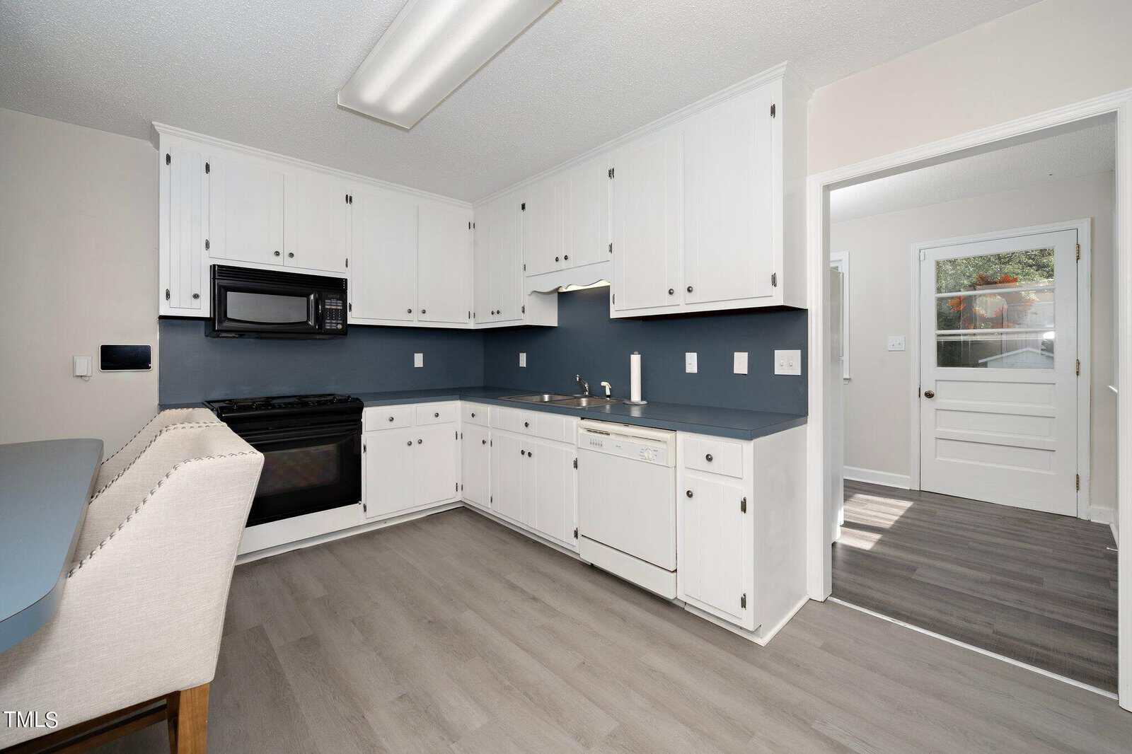 A kitchen with white cabinetry, a black microwave, and an oven. The countertops are blue, and there is a dishwasher. The floor is light gray wood, and there is a view of an adjacent room and a door leading outside.