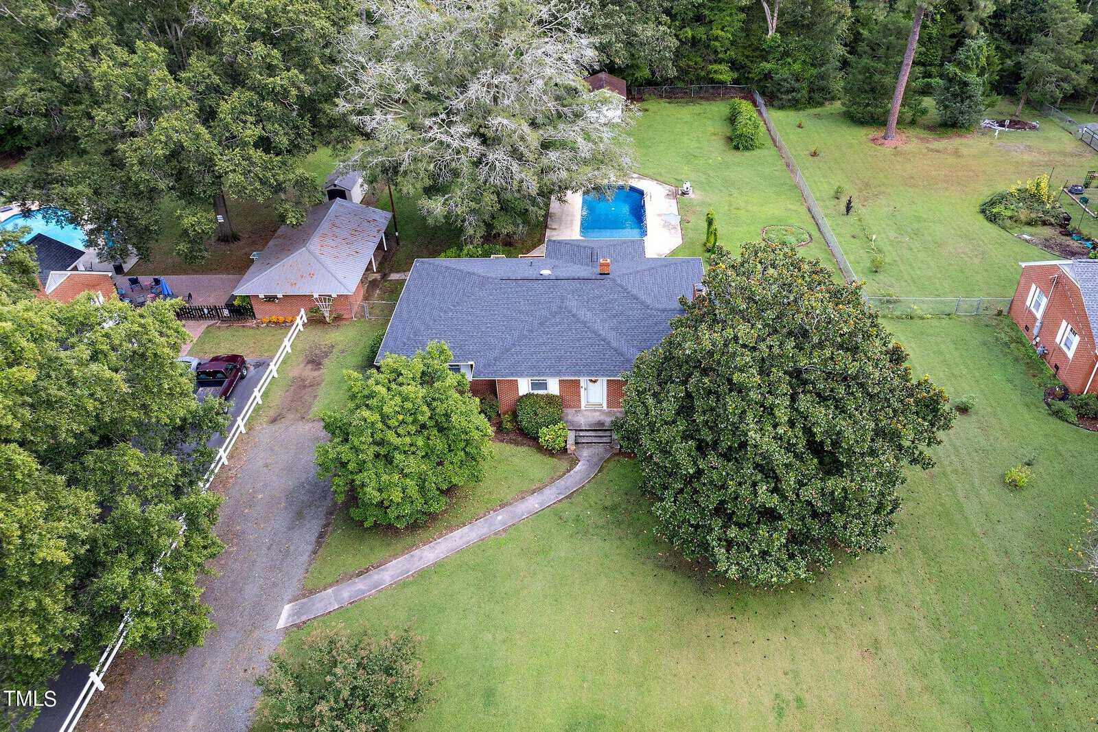 Aerial view of a single-story brick house at 604 18th Street, Butner, featuring a dark roof and surrounded by trees and a lush lawn. A swimming pool graces the backyard. The driveway elegantly curves toward the house, with a charming smaller building nearby in this verdant haven.