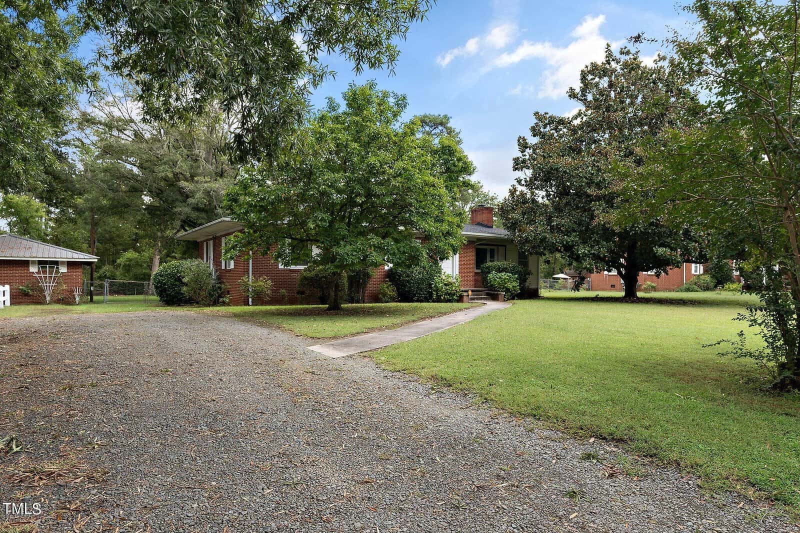 A brick house is surrounded by trees and a grassy lawn. A gravel driveway curves to the entrance, and another house is visible in the background. The sky is partly cloudy.