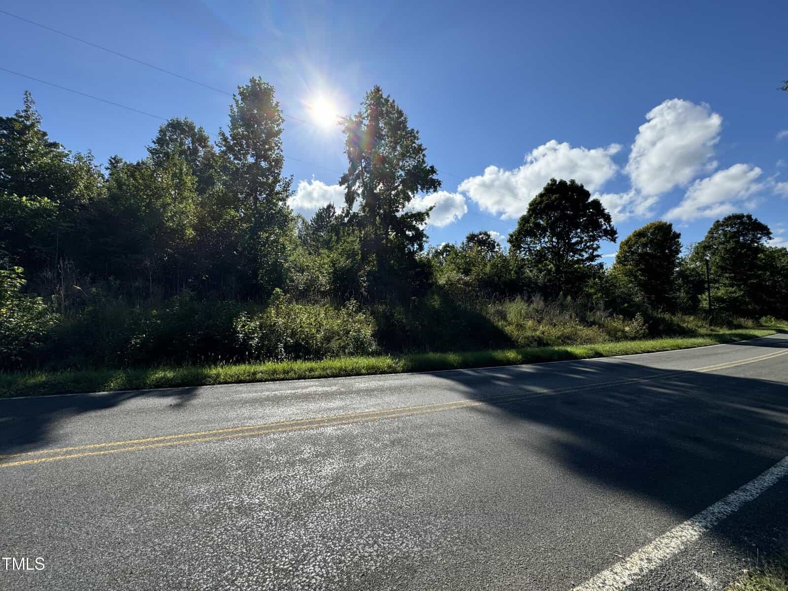 A sunlit road at 7635 Sam Hall Road in Oxford, bordered by lush greenery and tall trees under a bright blue sky with scattered clouds. Sunlight casts shadows on the road, creating a tranquil, countryside atmosphere.