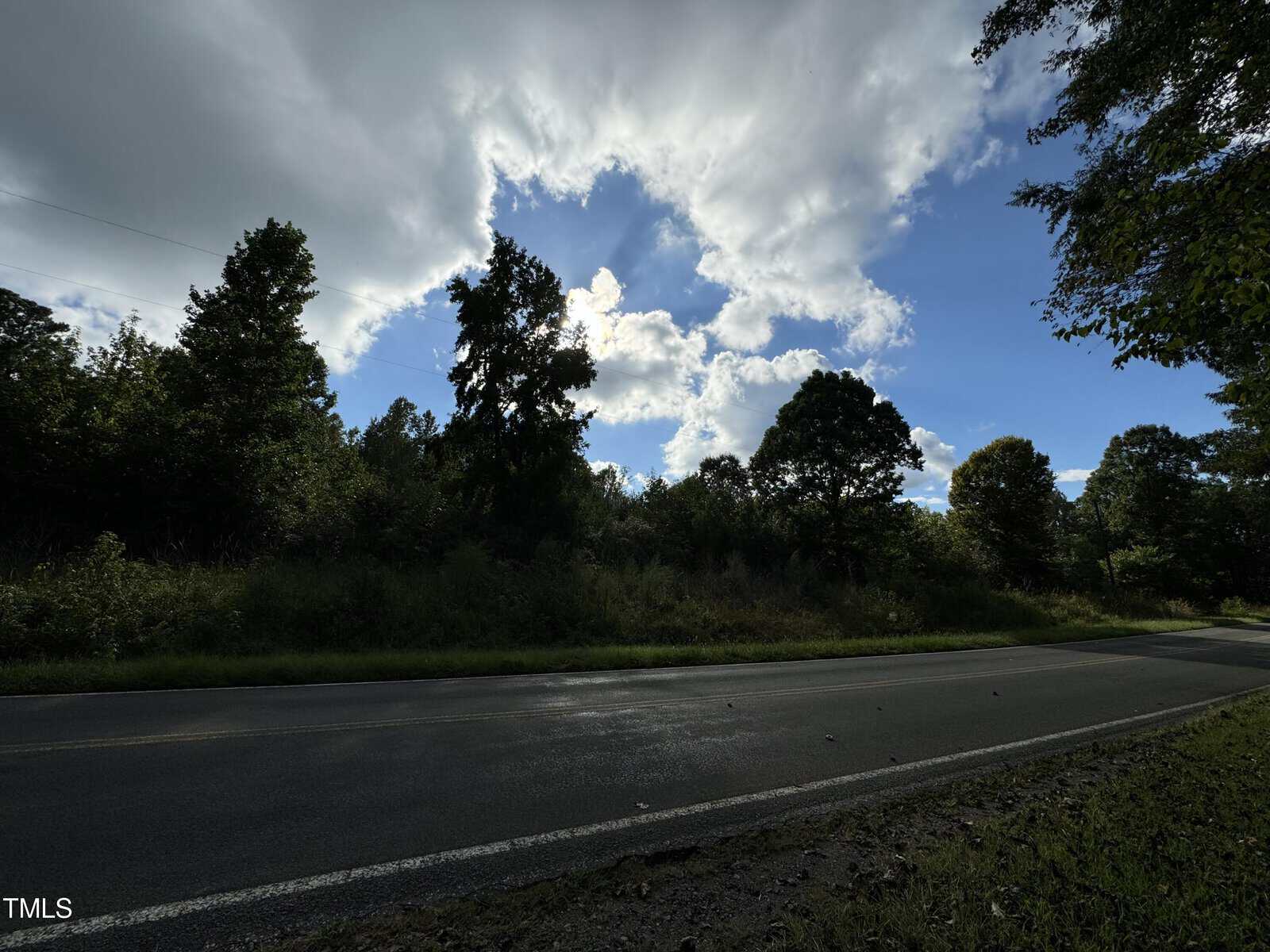 A scenic country road bordered by lush green trees under a partly cloudy sky. Sunlight breaks through the clouds, casting varying shades of light on the landscape. The road is empty, enhancing the tranquility of the scene.