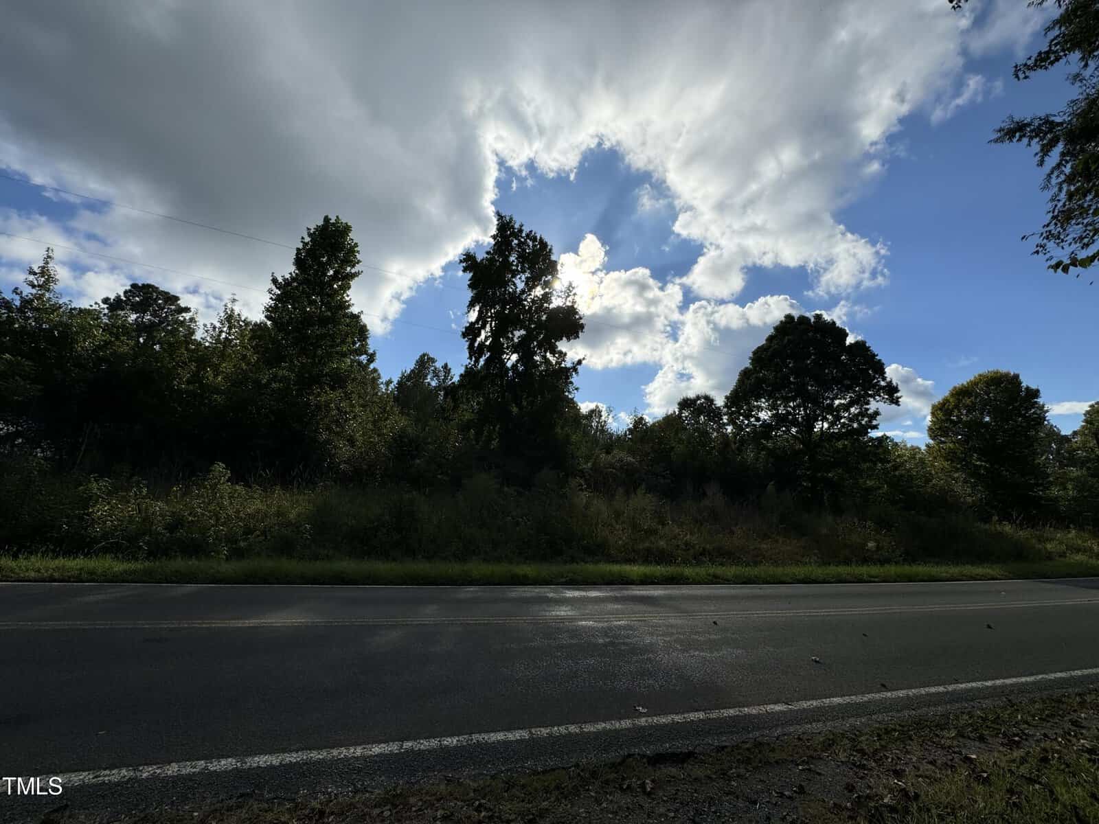 A road runs alongside a lush forest under a partly cloudy sky. Sunlight filters through the clouds, creating a serene atmosphere. Trees line the roadside, and the scene is calm and picturesque.