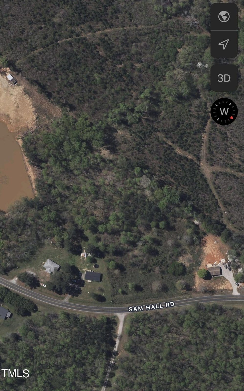 Aerial view of a rural landscape featuring a winding road labeled "Sam Hall Rd," surrounded by dense green trees and vegetation. A few buildings are visible near the road. A body of water is seen in the upper left corner.