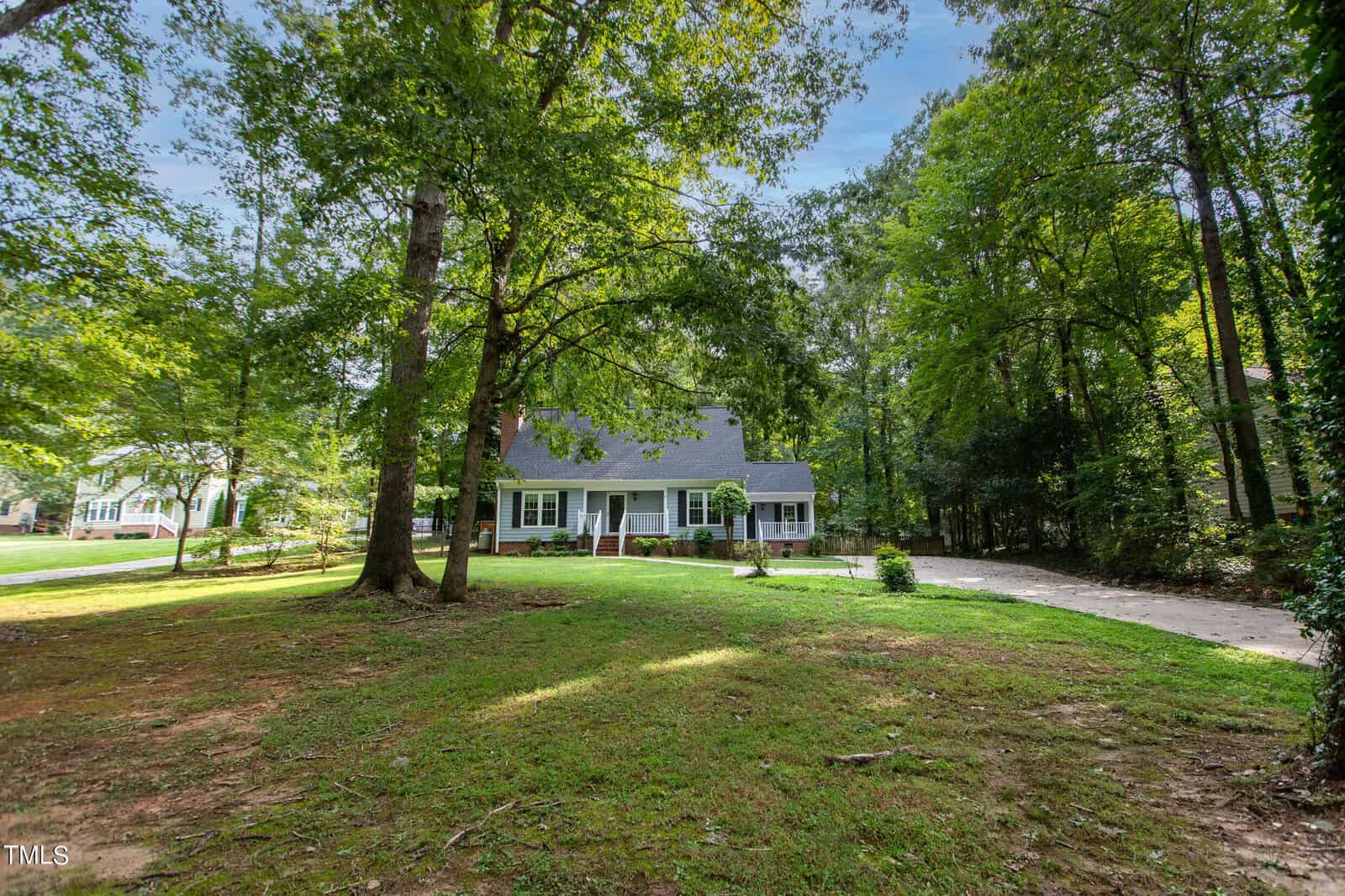 A charming white house with dark shutters and a steep roof sits surrounded by tall, leafy trees. The front yard is spacious, with a lush lawn and a long driveway to the right, leading to a side entrance. Sunlight filters through the trees, casting serene shadows.