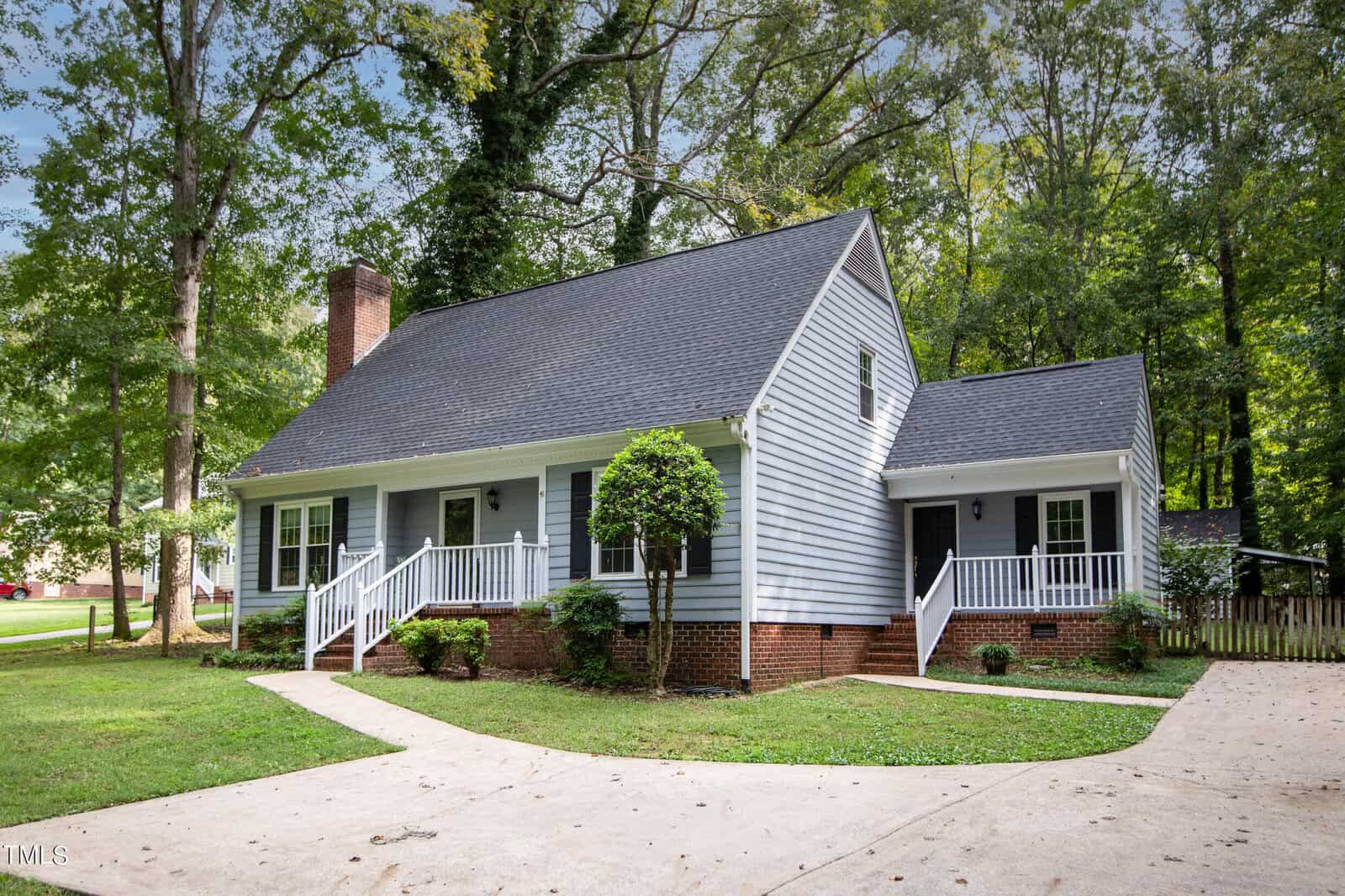 A charming two-story house at 201 S Stratford Drive with light gray siding and a dark gray roof, featuring a large white porch and railings. It includes a red brick foundation, green trees surrounding the property, and a curved driveway in the foreground in Henderson.