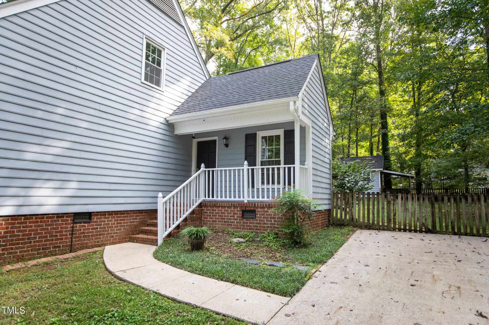 A small, quaint house with light blue siding, red brick foundation, and a white front porch. The house has a black front door, white railing, and is surrounded by trees. A curved concrete pathway leads from the driveway to the porch.