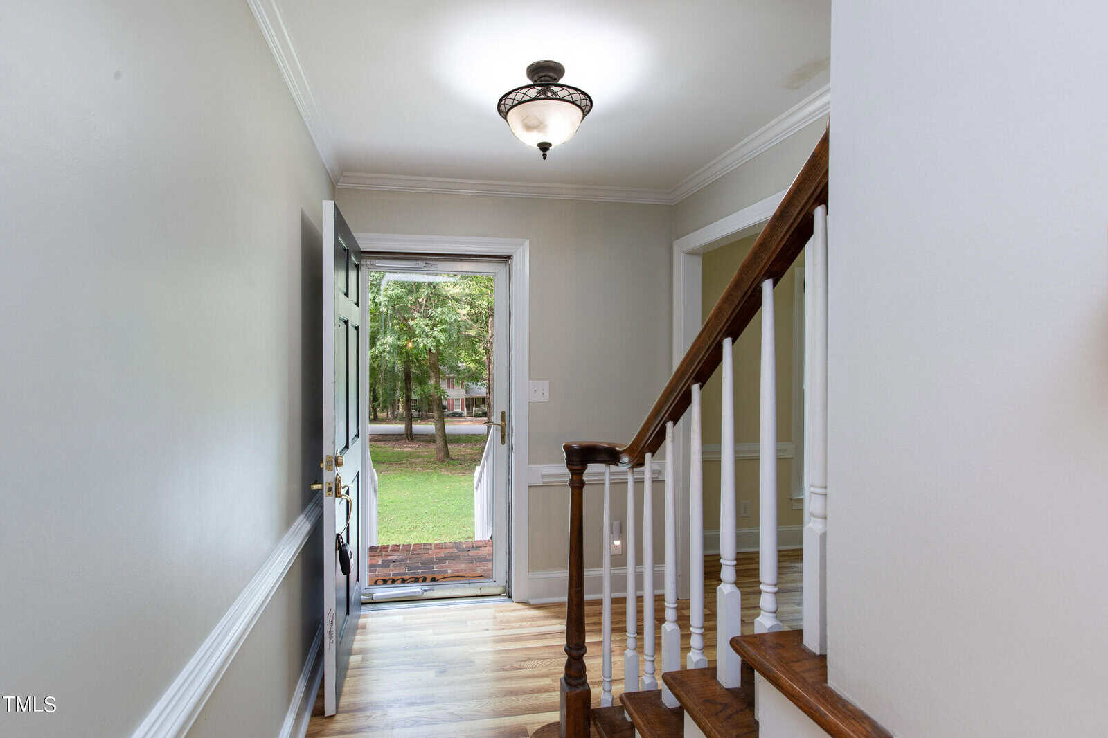 A well-lit entryway of a home featuring a staircase with wooden steps and white railings on the right. The front door, which is open, reveals a view of a green lawn and trees outside. The walls are painted light gray, and there is a light fixture on the ceiling.