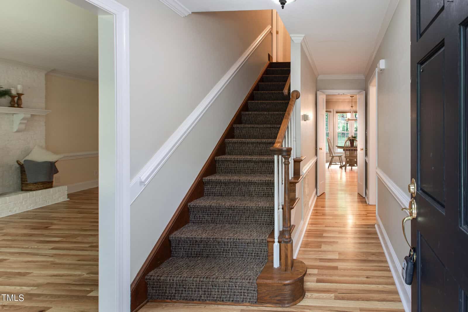 A hallway with a wooden staircase covered in patterned carpet leads to an upper floor. The walls are white with wainscoting, and the floor is hardwood. A room with a fireplace is visible on the left, and a dining area with a table and chairs is seen at the end of the hall.