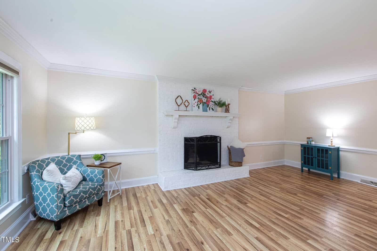 A bright living room with light wood flooring, a white brick fireplace, a floral painting above the mantel, and a blue-patterned armchair beside a small table with a lamp. A blue console table with decor stands against the wall, and large windows let in natural light.