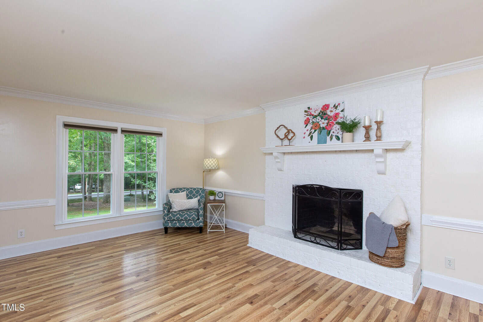 A bright living room with light-colored walls and wooden flooring. The room features a white brick fireplace with a decorated mantel, a patterned armchair next to a floor lamp and side table, and a large window allowing natural light to fill the space.