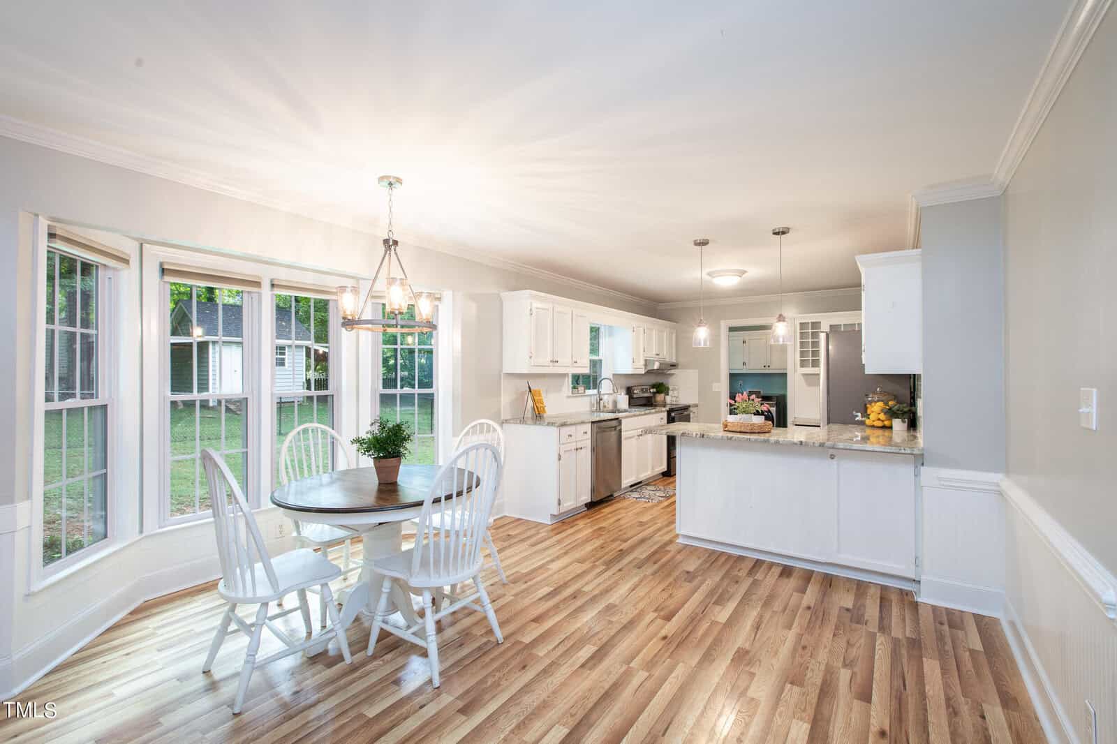 A bright kitchen and dining area with wooden flooring. The dining area features a round table with four white chairs by a large bay window. The kitchen has white cabinets, stainless steel appliances, and a central island with a fruit bowl and other decorations.