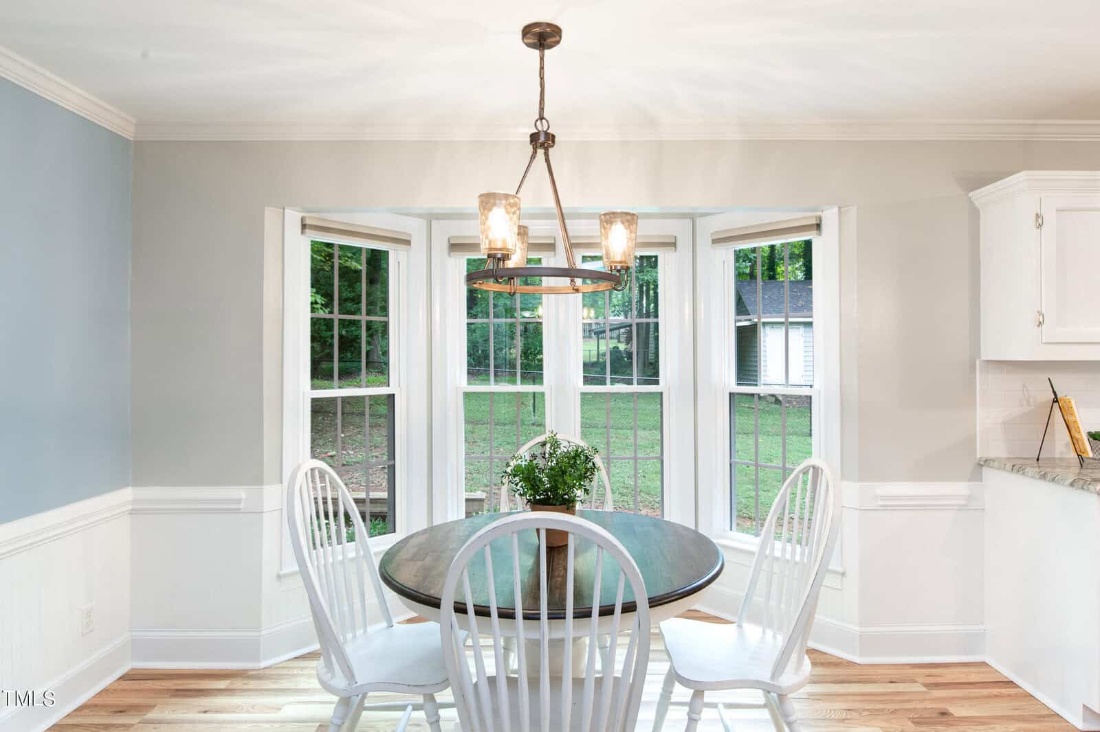 A cozy dining nook featuring a round wooden table with a small potted plant centerpiece and four white chairs. Natural light streams in from a bay window with views of a lush, green backyard. The walls are painted light gray, complemented by white trim and cabinets.
