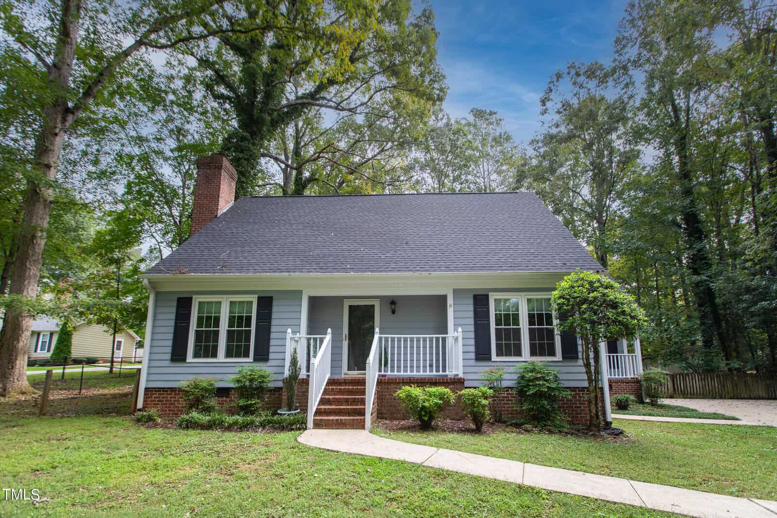 A charming white house with a dark roof, brick foundation, and blue shutters, nestled among tall trees. A curved path leads to the front steps, with a grassy lawn and a neatly trimmed hedge lining the entrance.