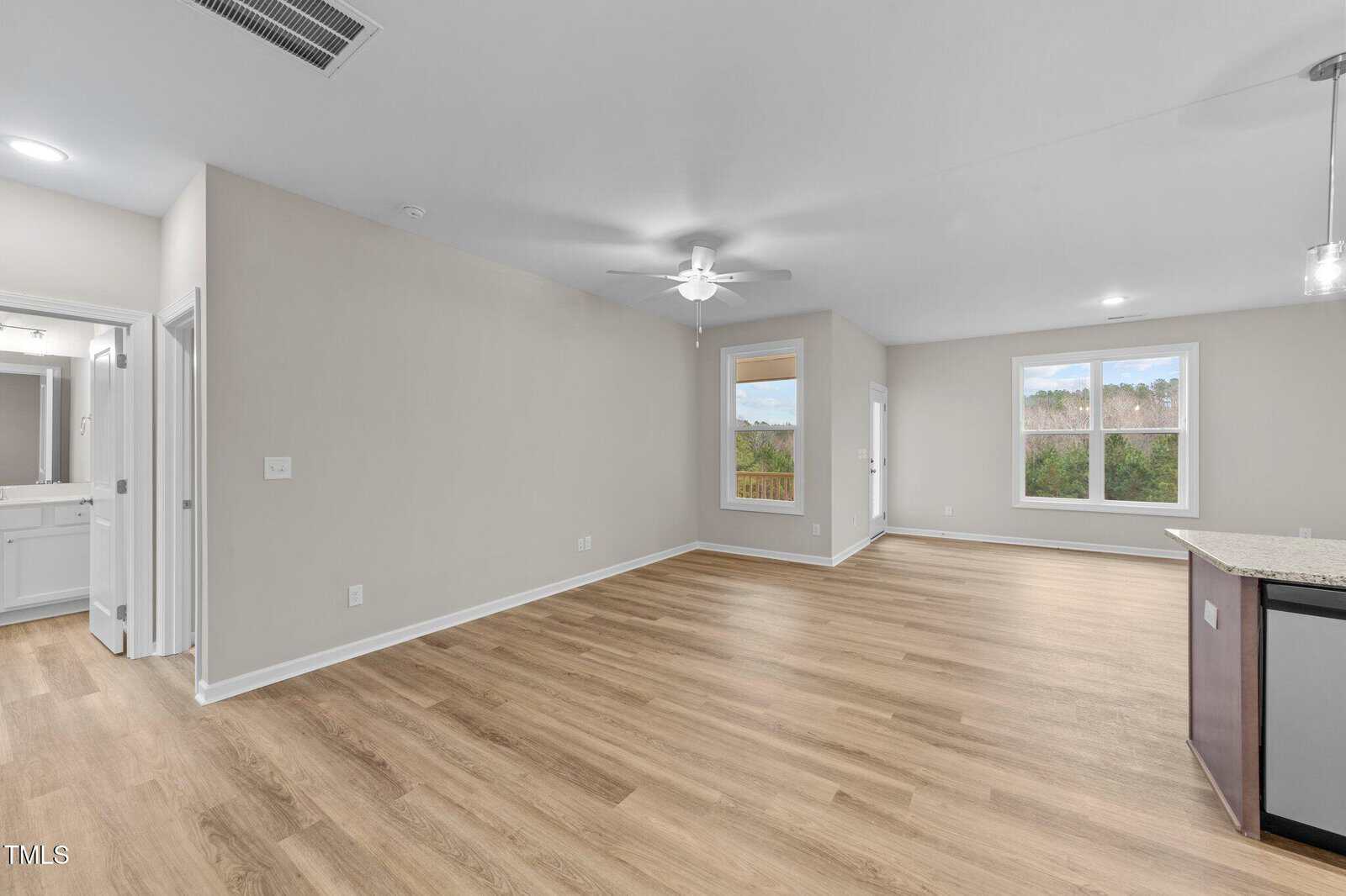 A spacious, empty living area with light wood floors, beige walls, ceiling fan, and large windows. The open layout connects to a kitchen with granite countertops. A bathroom is visible through an open door on the left.