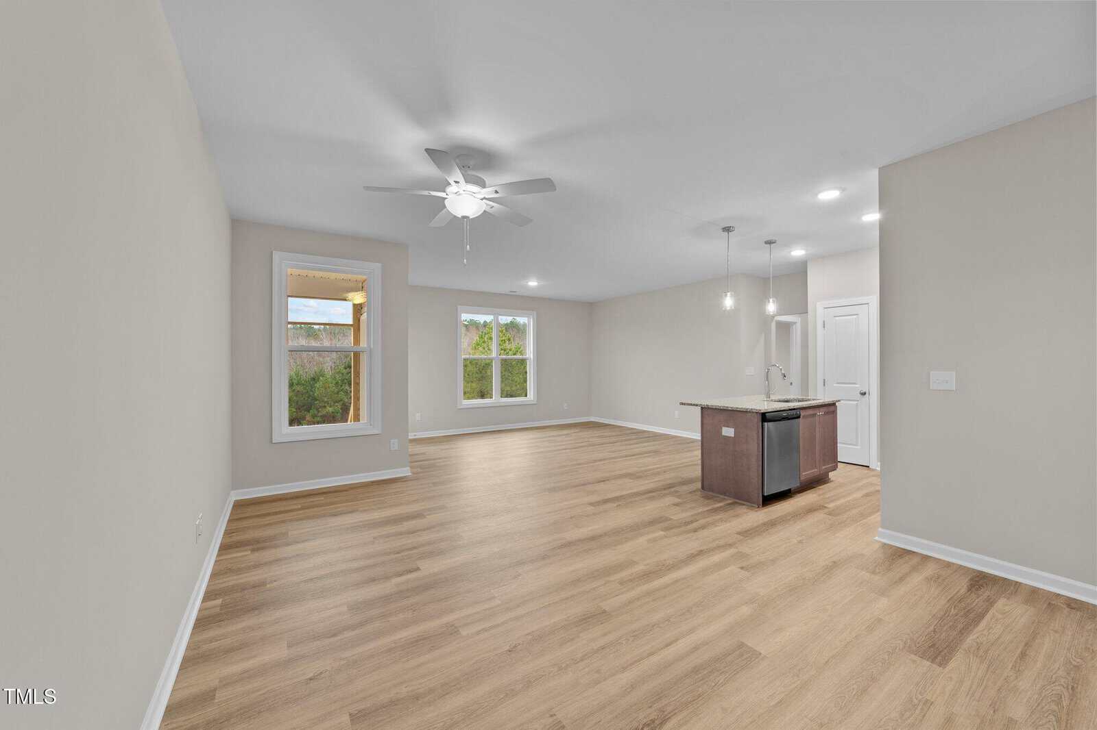 A spacious, unfurnished open-concept living area with light wood flooring, beige walls, large windows, a ceiling fan, and a kitchen with an island, pendant lights, and white door.