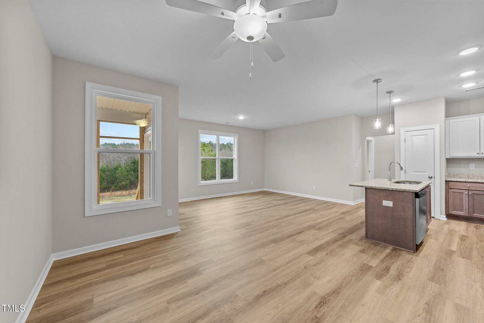 A spacious, empty open-concept living area with light wood flooring, neutral walls, large windows, ceiling fan, and a kitchen with an island and pendant lights. A covered porch is visible through the side window.