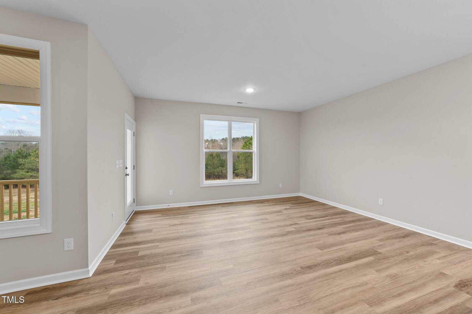 A spacious, empty room with light wood flooring, beige walls, a large window, a glass door leading outside, and white baseboards. The room is well-lit with natural light.