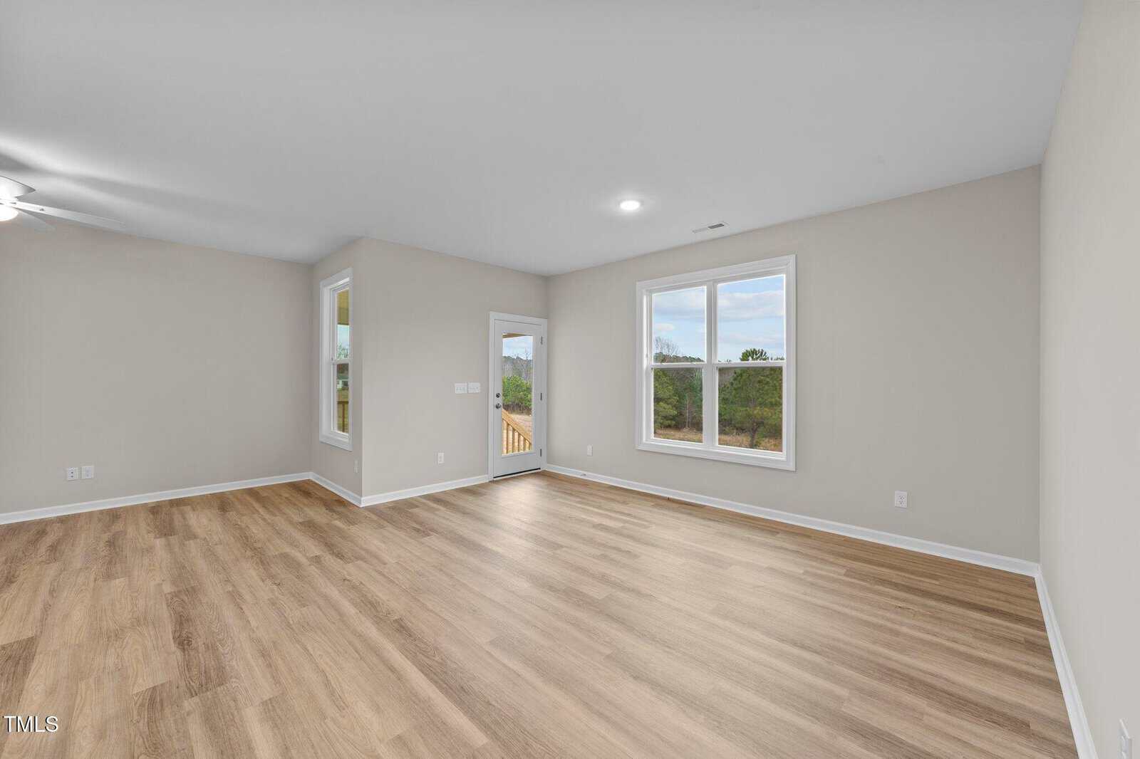 Empty room with light wood flooring, beige walls, two large windows, a glass door leading outside, and a ceiling fan in the back left corner. The room appears bright with natural light.