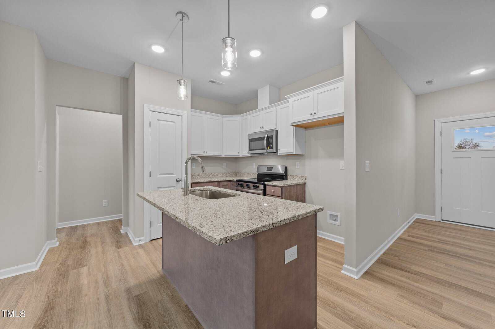 A modern kitchen with light beige walls, wood flooring, white cabinets, a granite countertop island with a sink, stainless steel appliances, and pendant lights hanging from the ceiling.