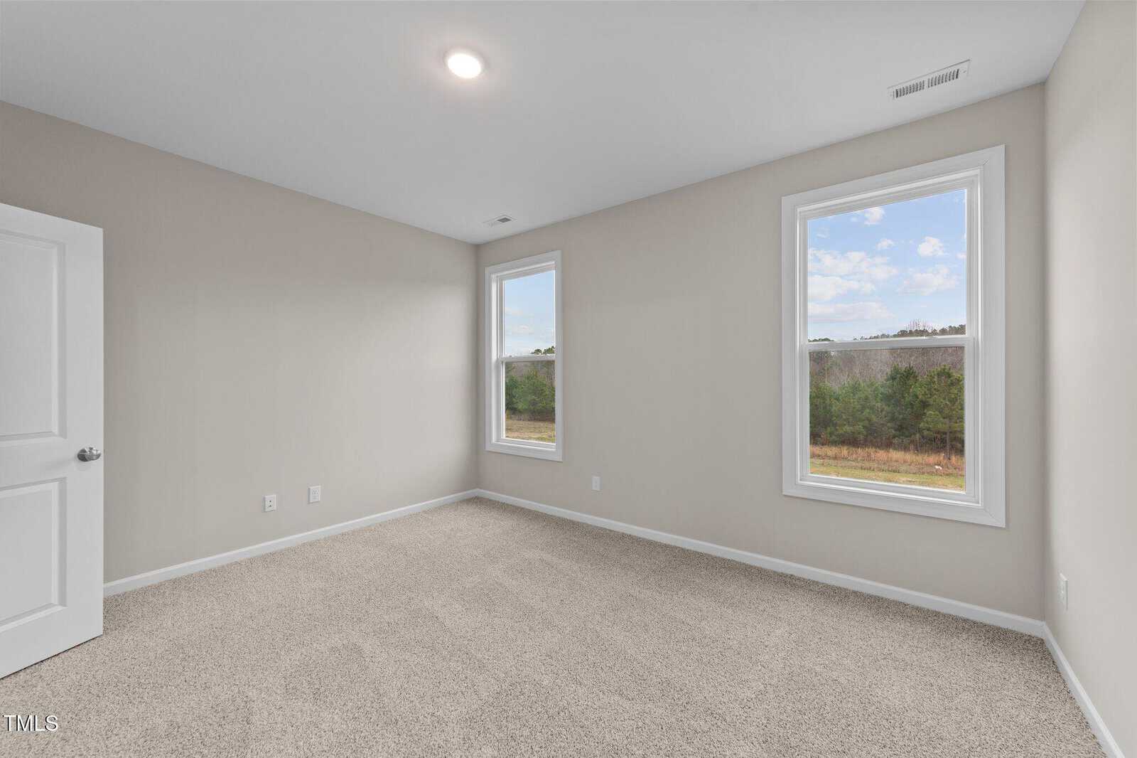 Empty room with beige carpet, light-colored walls, two large windows showing a view of trees and sky, a white ceiling light, and an open white door on the left wall.
