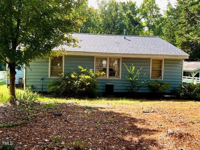 A small, single-story house with light blue siding, surrounded by trees and bushes. The yard has a mix of dirt and sparse grass. Large windows are visible on the house's front side, and the house has a dark gray roof. The background features dense, green foliage.