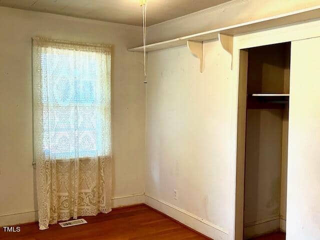 A small, empty room with a single window covered by lace curtains. The walls are white and the hardwood floor is light-colored. To the right, there is an open closet with a wooden rod, and a narrow shelf runs along the wall above the closet.
