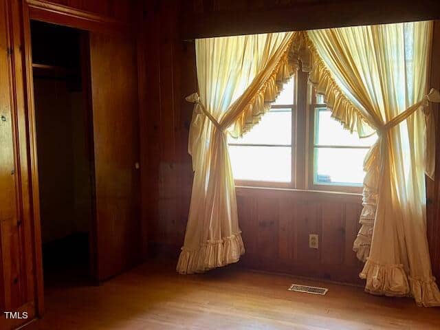 A room with wooden walls and floor features a window with ruffled cream curtains tied back on each side. The room has an open doorway leading to a closet with no door, revealing darkness inside. An air vent is visible on the floor near the window.