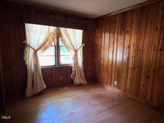 A small, empty room with wood-paneled walls and a wooden floor. A lone window with sheer, ruffled curtains letting in natural light is centered on one wall. The room appears to be well-maintained and simple, with no furniture or decorations.