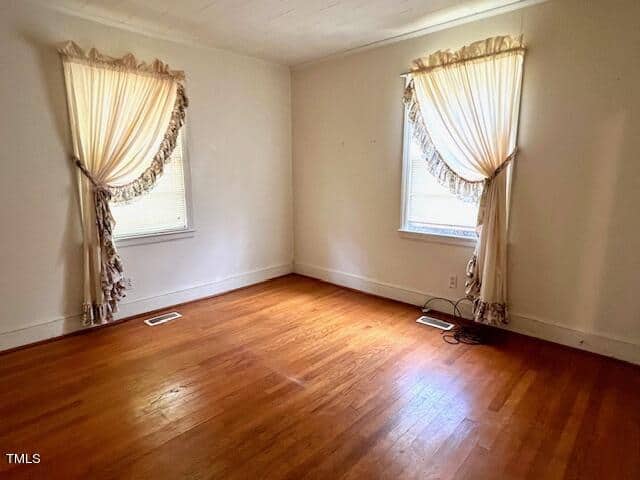A vacant room with light-colored walls, two windows covered by beige drapes, and hardwood floors. Sunlight softly enters the space, making it appear warm and inviting.