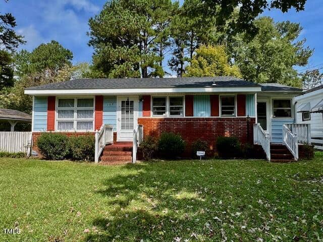 A quaint, single-story house at 134 W Parker Street in Henderson, with red brick and light blue siding, featuring a white door with red shutters. The home has two sets of steps leading to the front entrances and is surrounded by a well-maintained lawn, bushes, and large trees in the background.