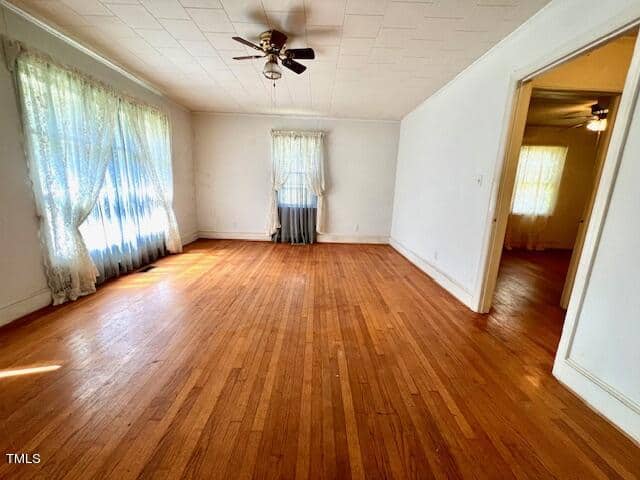 A spacious, empty room with hardwood floors and white walls. Daylight filters in through lace curtains over two windows. A ceiling fan is mounted centrally on the white ceiling. A doorway on the right leads to another room with a similar setup.
