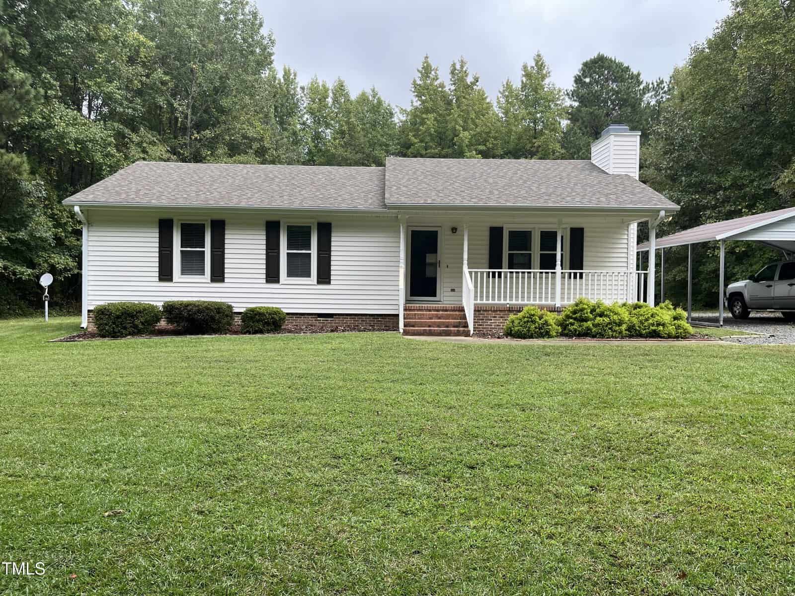 This charming single-story white house on 3143 Hester Road in Creedmoor features a gray roof and a welcoming front porch with steps leading up. The lawn is neatly cut, complemented by bushes along the front. A car is partially visible under the carport to the right, with trees as a serene backdrop.