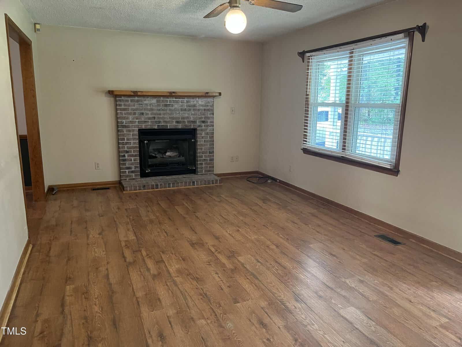 A spacious, unfurnished living room with wooden floors and a ceiling fan. A brick fireplace with a wooden mantel is centered on one wall. Large windows with blinds allow natural light to fill the room.