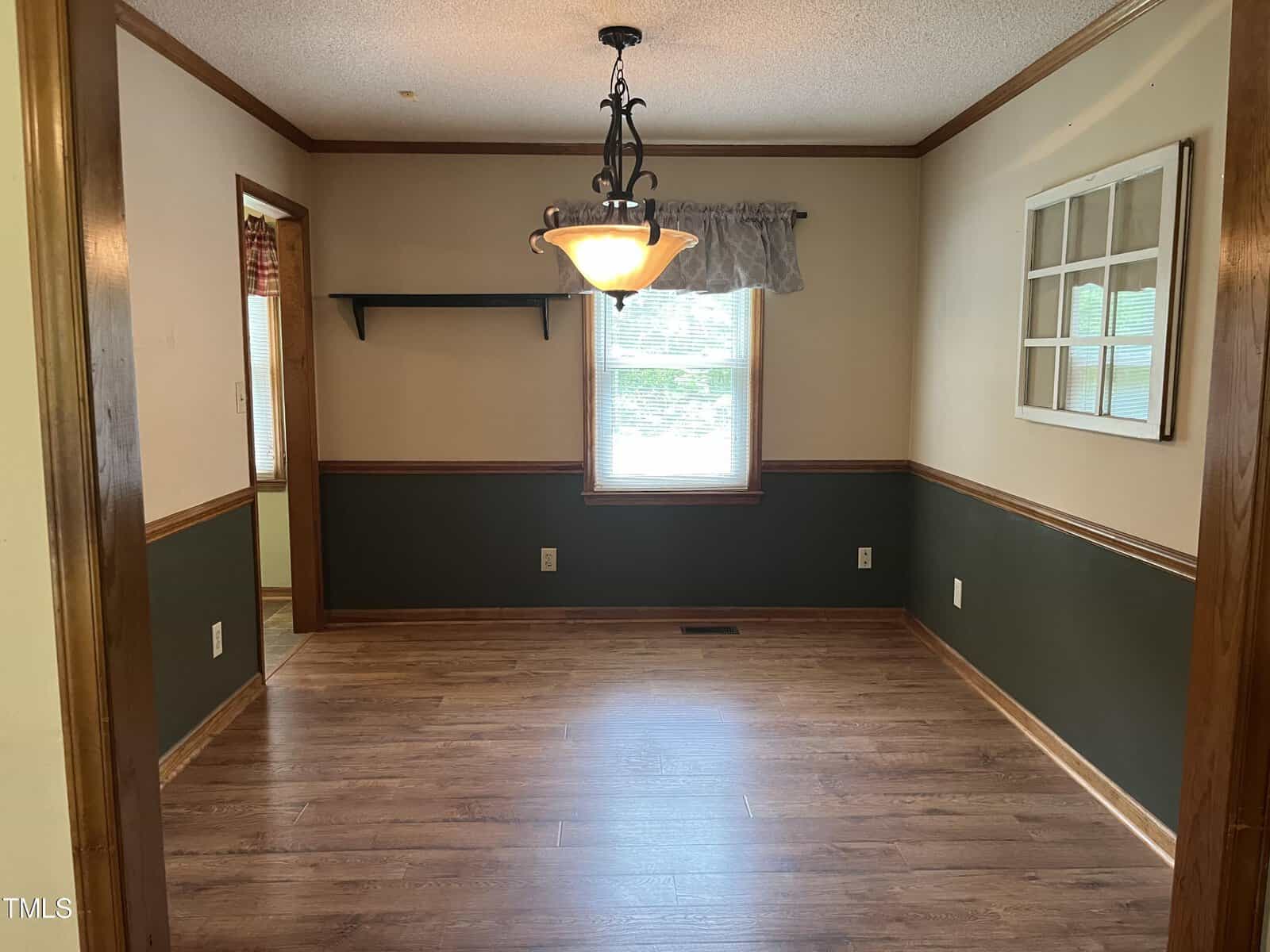 A vacant room with hardwood floors and two-tone walls, featuring a lower dark green section topped with beige. A window with a valance lets in natural light. A central pendant light hangs from the ceiling. An open doorway leads to another room.